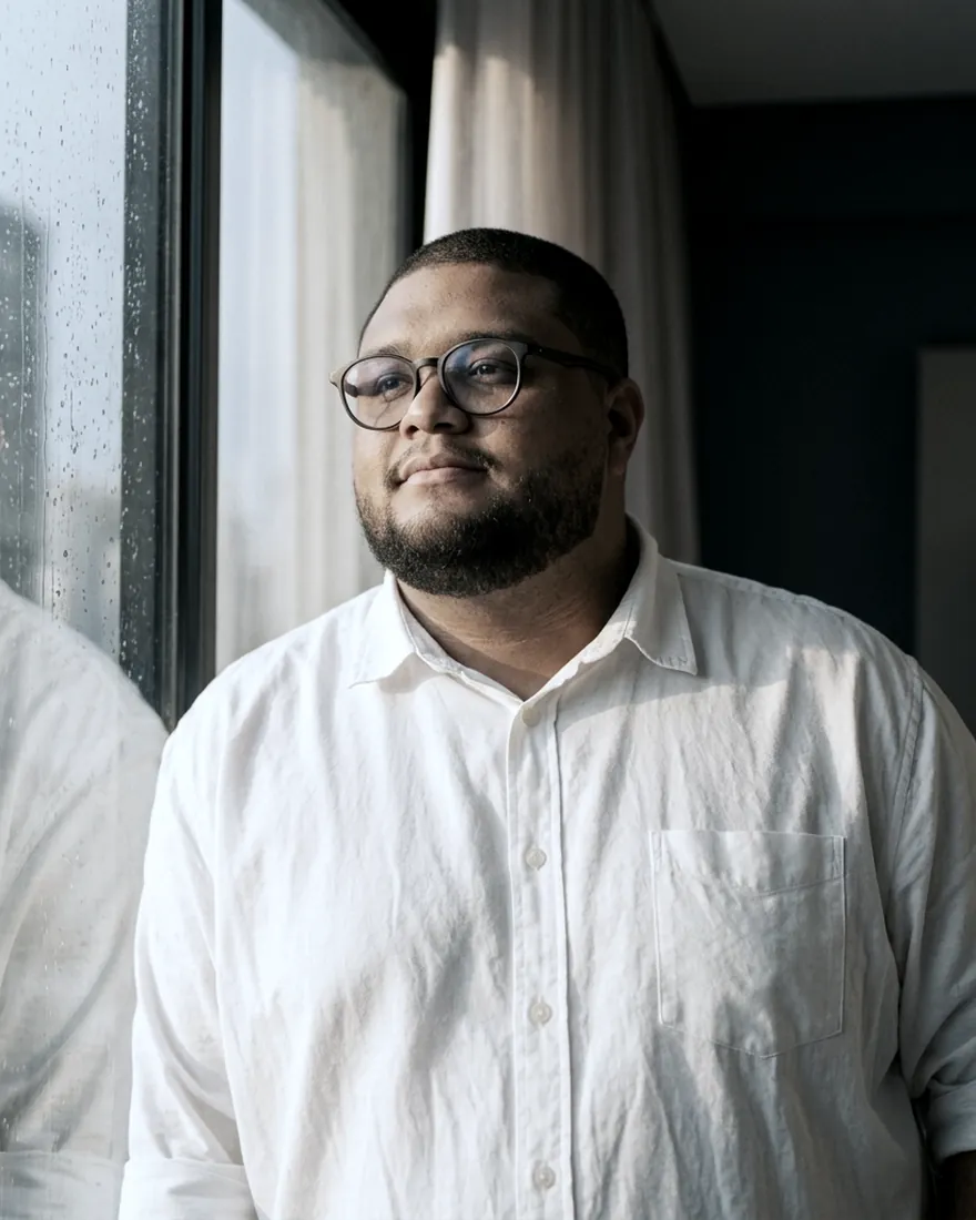 Matheus Dantas de camisa branca, junto a uma janela, em luz natural